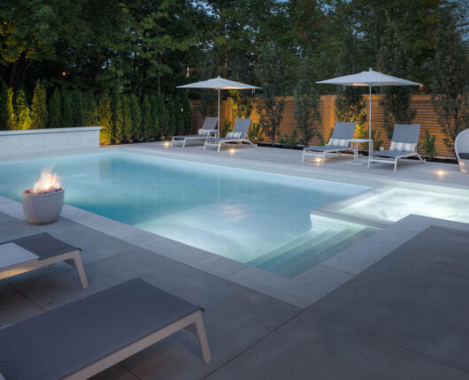 View of a residential backyard at dusk where a well-lit, modern concrete pool is shown with adjacent pool loungers and umbrellas, along with treed landscaping and large fire pots.