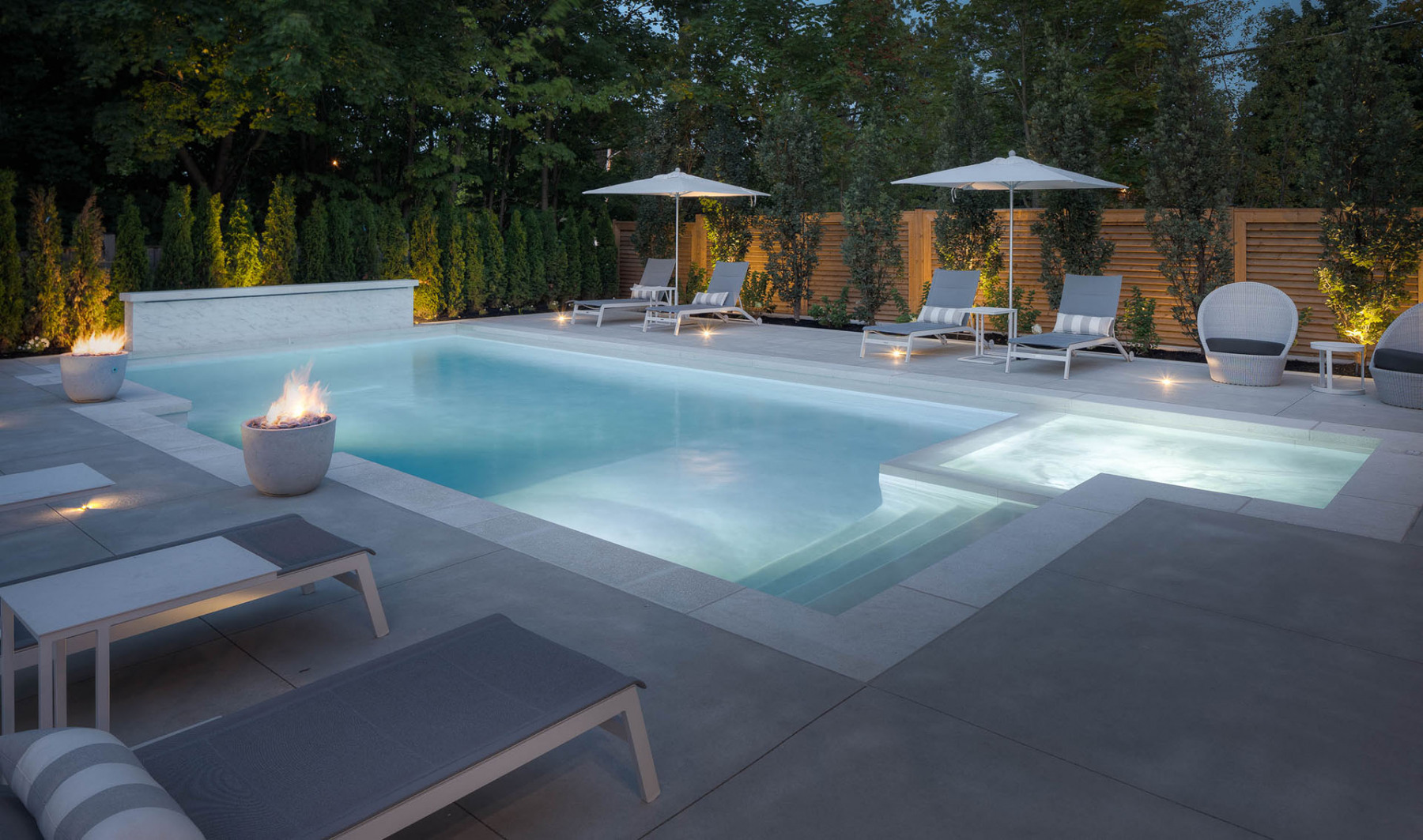 View of a residential backyard at dusk where a well-lit, modern concrete pool is shown with adjacent pool loungers and umbrellas, along with treed landscaping and large fire pots.