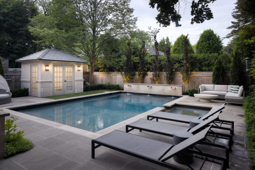 Three lounge chairs, a seating area and a small pool house surround a modern concrete pool in a residential backyard.
