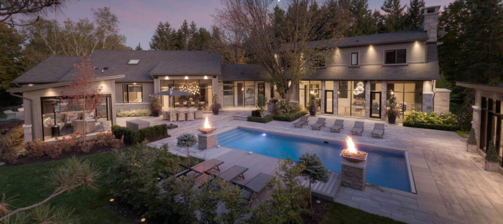 View of a large residential backyard at dusk, featuring a mid-sized concrete pool with two fire columns, lounge chairs and seating area.