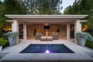 View of a outdoor lounge area with warm lighting, a small pool, tv, fireplace and seating surrounded by lush greenery.