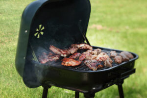 Portable barbecue with steak and pork grilling outdoors; grass in the background.