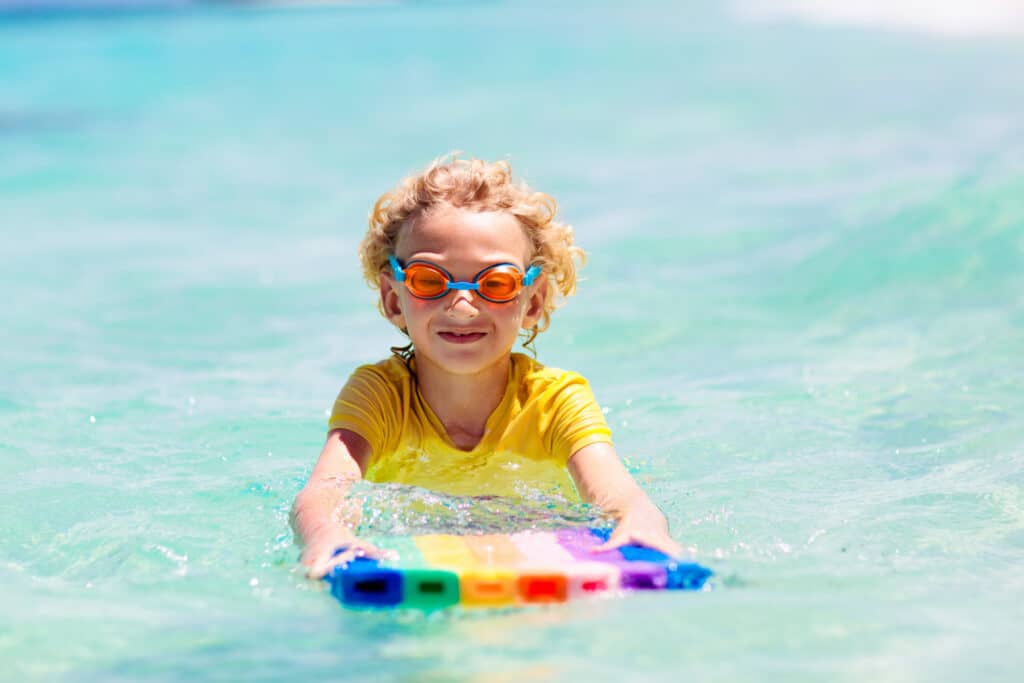 A young child in a bright yellow swim shirt and goggles swims in the ocean with a rainbow-coloured body board.