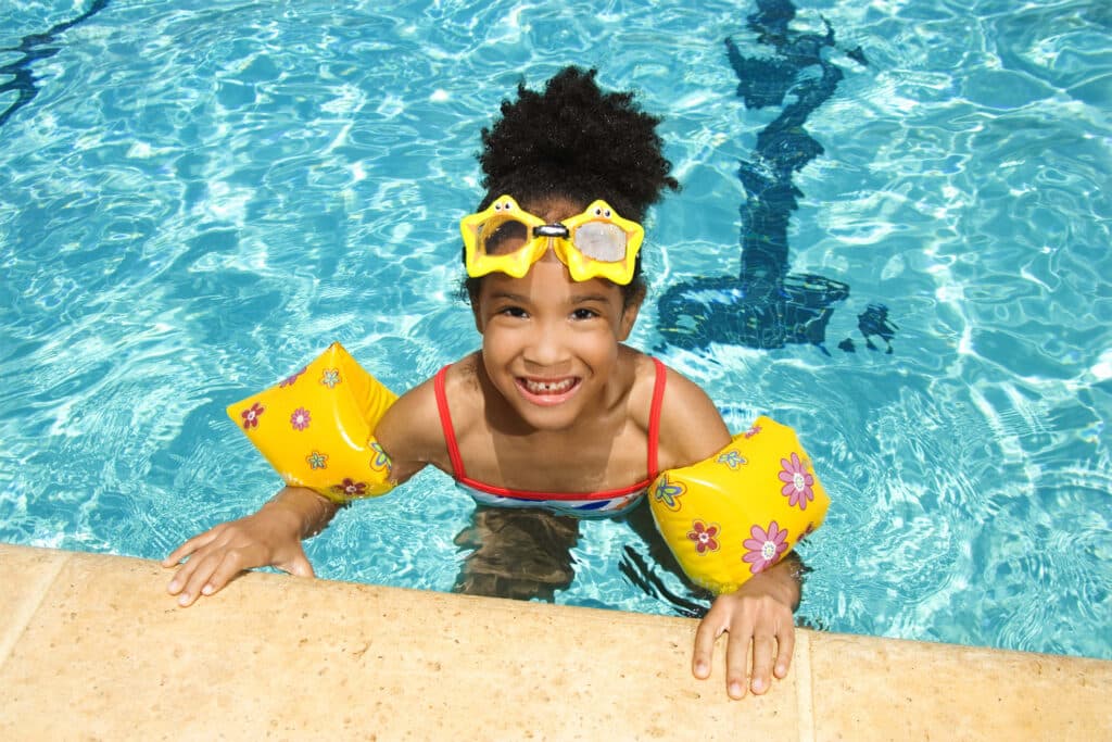 A young girl wearing yellow arm floaties and star-shaped goggles smiles at the camera from the edge of a swimming pool.