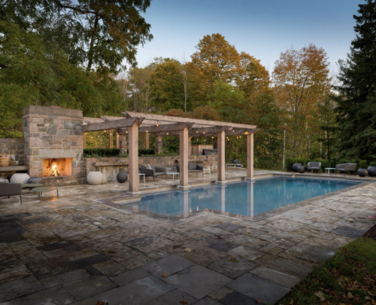 A custom concrete pool is shown with a large stone fireplace, pergola and lounge area during a beautiful fall day, with changing tree colours and fallen leaves.