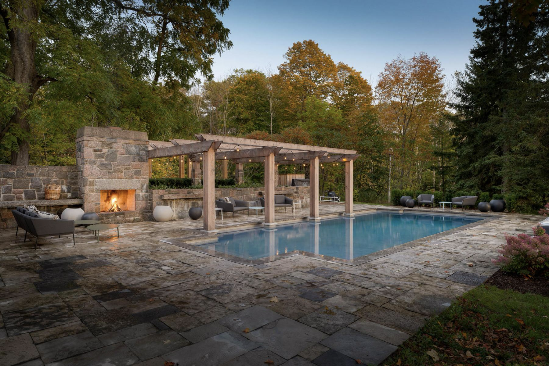 A custom concrete pool is shown with a large stone fireplace, pergola and lounge area during a beautiful fall day, with changing tree colours and fallen leaves.