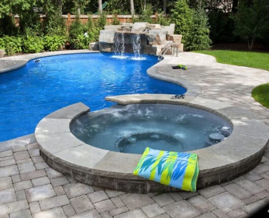 Alt text: A custom vinyl pool and attached hot tub surrounded by grey interlocking stone with a rock waterfall in a sunny residential backyard