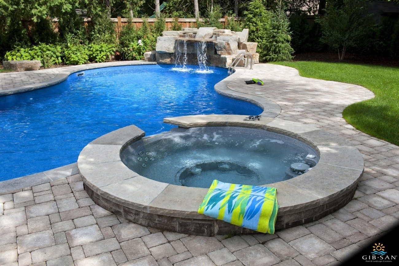 Alt text: A custom vinyl pool and attached hot tub surrounded by grey interlocking stone with a rock waterfall in a sunny residential backyard