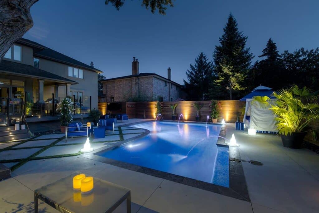 Evening shot of a concrete pool in a residential backyard surrounded by landscape lighting, water features and a lounge area.