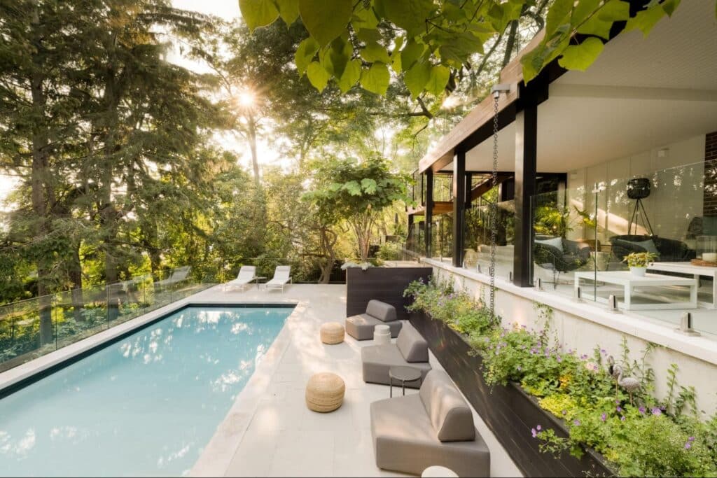 A stunning view of a concrete pool in a backyard at sunset surrounded by various greenery and trees along with multiple lounge areas.