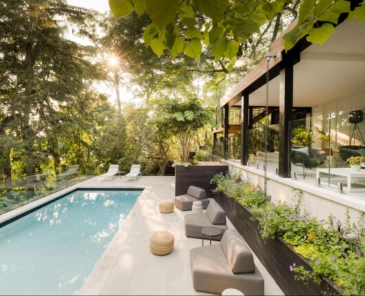 A stunning view of a concrete pool in a backyard at sunset surrounded by various greenery and trees along with multiple lounge areas.