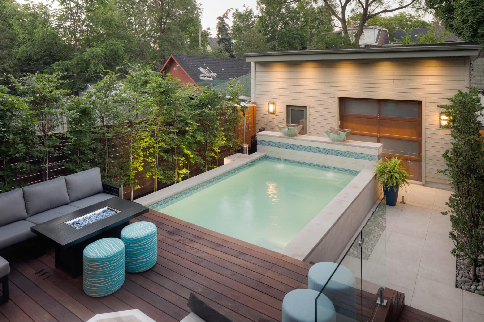 A swim spa encased in stone featuring two small water features is shown in a small, lush residential backyard between a modern garage unit and a lounge area with seating.