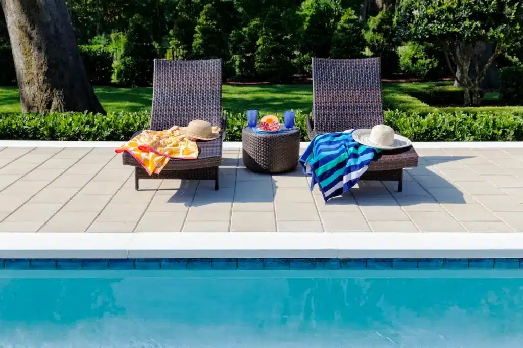 Inground pool edge with light-coloured pool coping and patio pavers, two lounge chairs with towels and hats, and a small table with drinks in a landscaped backyard.