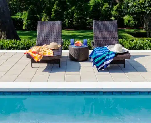 Inground pool edge with light-coloured pool coping and patio pavers, two lounge chairs with towels and hats, and a small table with drinks in a landscaped backyard.