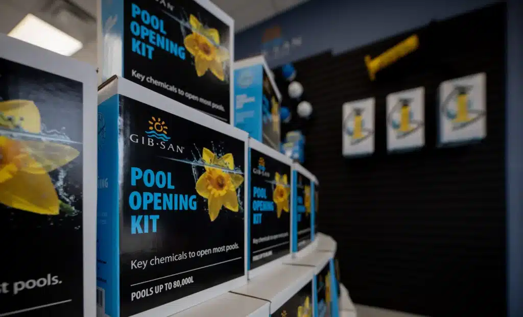 Row of Gib-San Pool Opening Kit boxes displayed on shelves in a pool supply store, with additional products and signage blurred in the background.