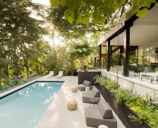 Modern backyard pool beside a contemporary home with glass railing, outdoor lounge seating, and lush landscaping in warm evening light.