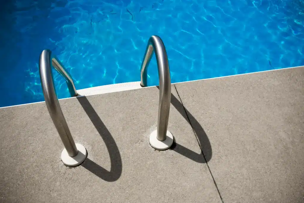 Metal pool ladder installed on concrete deck beside clear blue swimming pool water during spring pool opening preparation