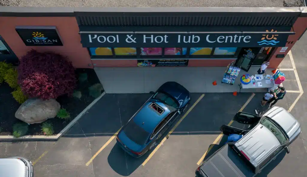 Aerial view of the Gib-San Pool & Hot Tub Centre storefront with cars in the parking lot and people gathered near a display table outside.