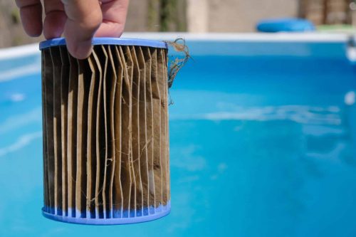 A dirty replacement pool filter cartridge is held in a person’s hand with the pool in the background.